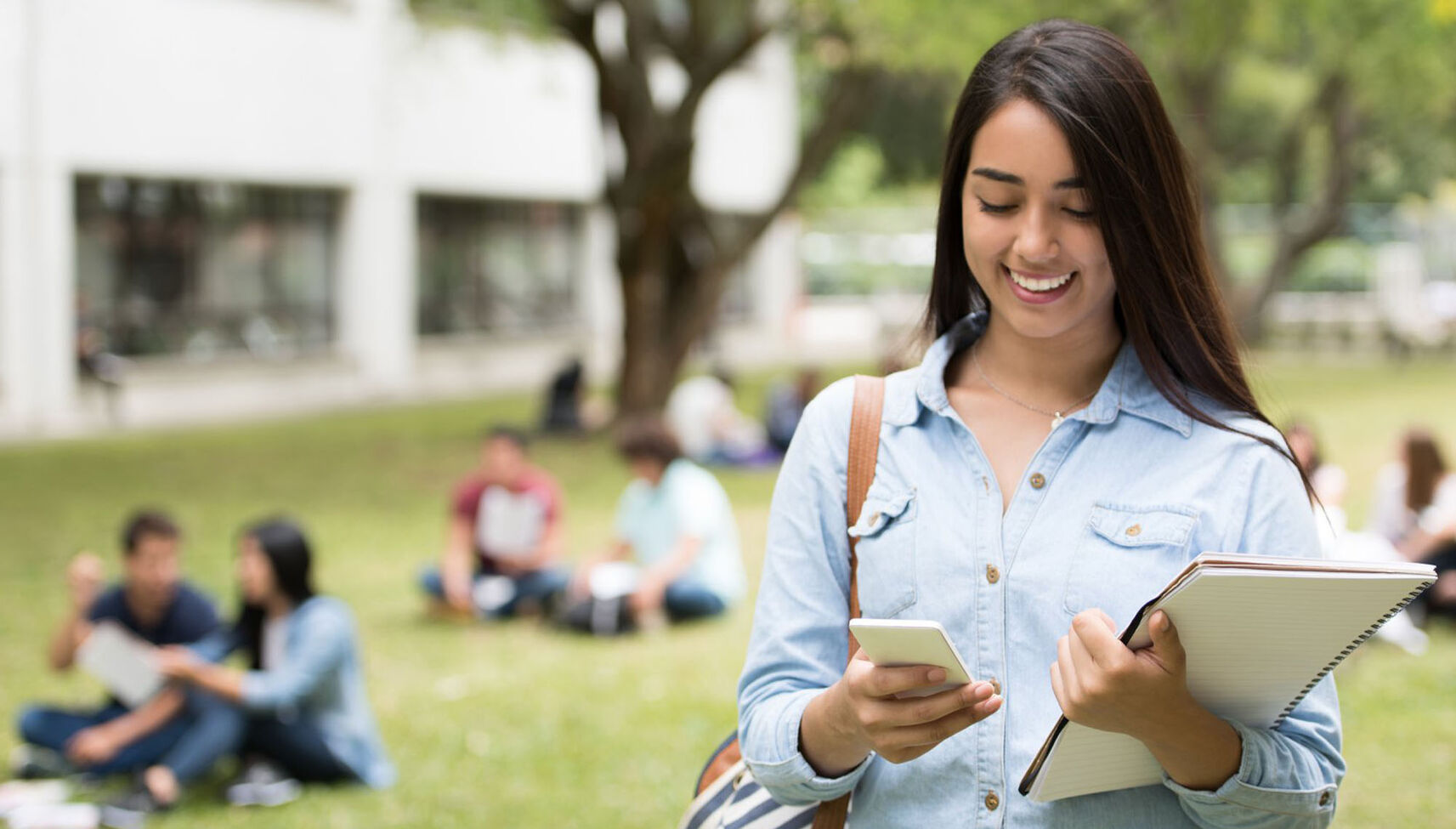 A female student walking outdoors holding her books and looking at her phone