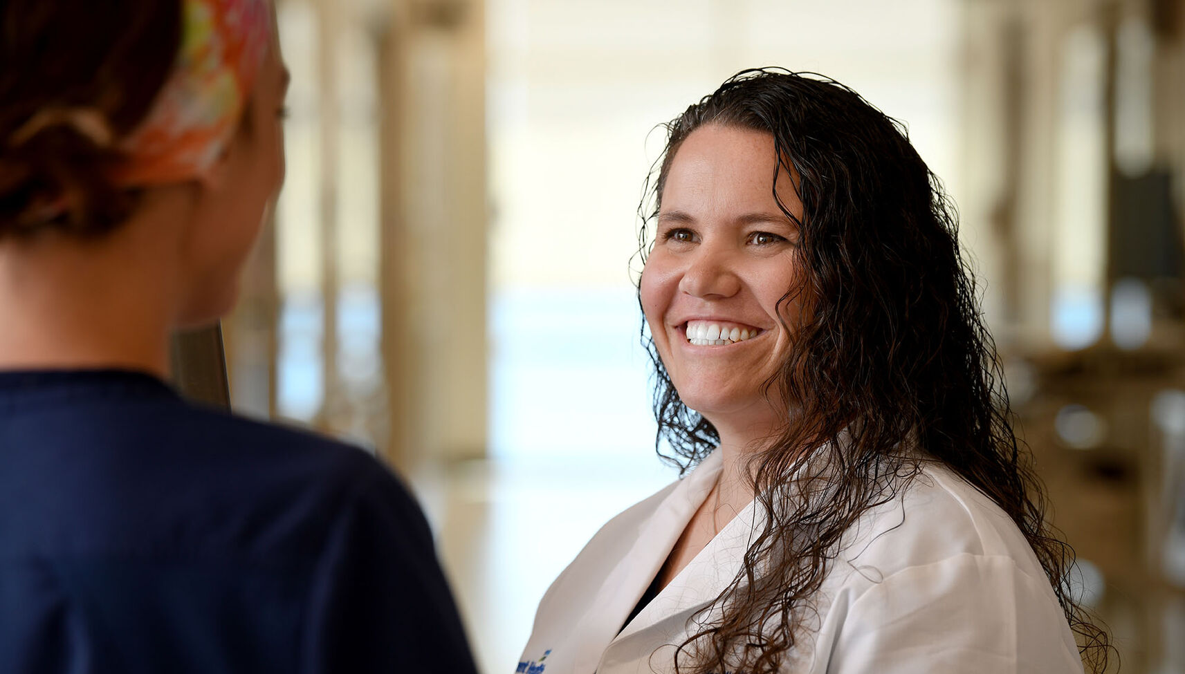 A nurse in a white coat smiling and talking with another nurse