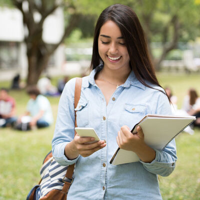 A female student walking outdoors holding her books and looking at her phone